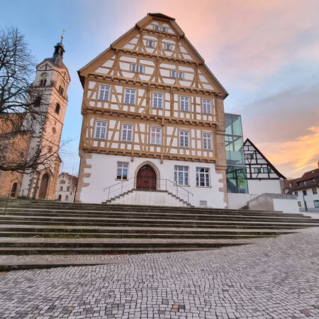 001_002 Ein historisches Fachwerkgebäude mit mehreren Fenstern steht neben einem Turm, beide umgeben von gepflasterten Stufen und einem Himmel im Sonnenuntergang.