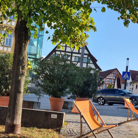 001_004 Auf dem Bild sieht man eine ruhige Straßenszene mit Fachwerkhäusern im Hintergrund, einem Baum, mehreren Pflanzentöpfen und orangenen Liegestühlen im Vordergrund.