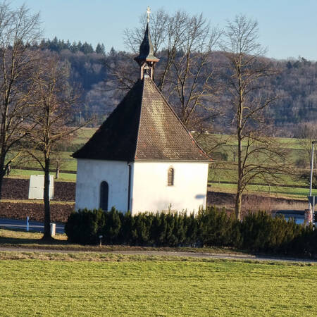 001_005 Eine kleine, weiße Kapelle mit einem steilen, dunklen Dach steht umgeben von Bäumen und Wiesen in einer ländlichen Landschaft.