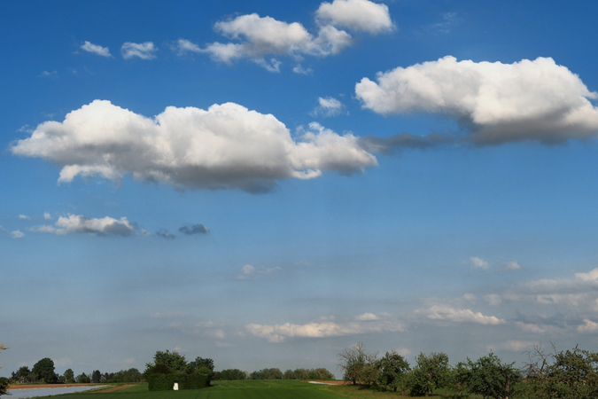 002_001 Eine grüne Wiese mit Bäumen im Vordergrund und großen weißen Wolken an einem blauen Himmel.