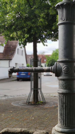 002_002 Ein alter, metallener Wasserbrunnen steht auf einem Bürgersteig vor einem Baum und Wohnhäusern im Hintergrund.