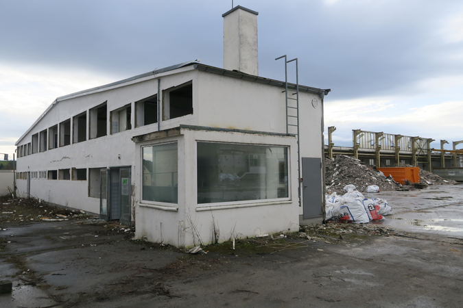 Ein verlassenes, zweistöckiges Gebäude steht auf einem unordentlichen Gelände mit Schutt und grauem Himmel im Hintergrund.