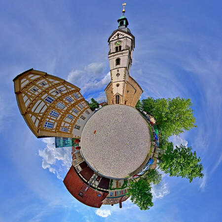 004_003 Eine Miniaturwelt-Darstellung zeigt eine gepflasterte Fläche in der Mitte, umgeben von Fachwerkhäusern, einem Kirchturm und grünen Bäumen unter einem blauen Himmel.