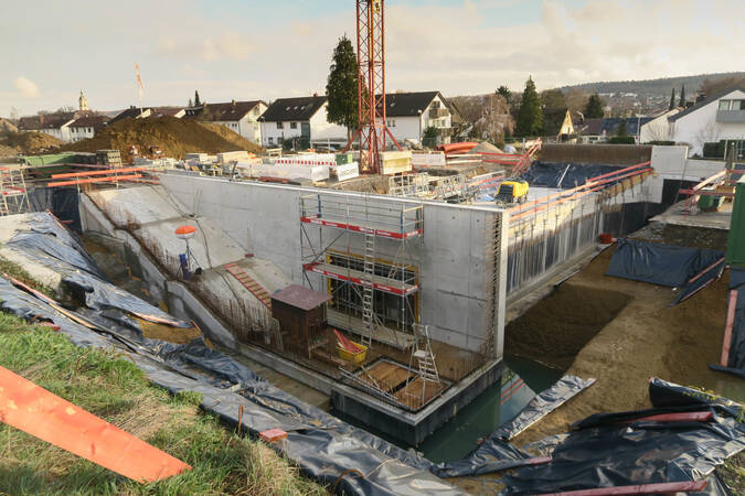 005_005 Eine Baustelle mit tiefen Baugruben, teilweise errichteten Betonwänden, Baugerüsten und einem Kran in einer Wohngegend.