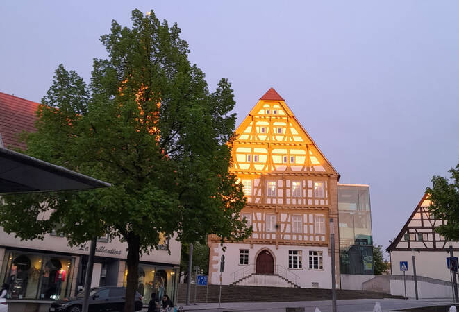 005_002 Ein Fachwerkhaus mit beleuchtetem Giebel steht neben einem Baum auf einem Platz, während die Abenddämmerung den Himmel erhellt.
