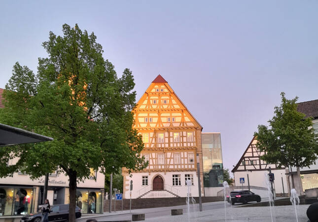 005_004 Ein Fachwerkhaus mit beleuchtetem Giebel steht neben einem Baum auf einem Platz, während die Abenddämmerung den Himmel erhellt.