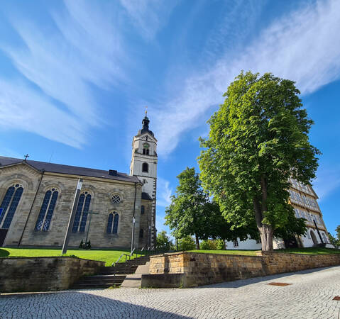 007_001 Eine Kirche mit einem Turm steht neben einem großen, grünen Baum unter einem klaren blauen Himmel.