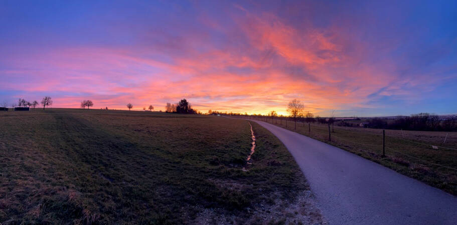 008_002 Eine Landschaft mit einer Wiese und einem Weg, die in einen farbenfrohen Sonnenuntergang tauchen, der den Himmel in leuchtenden Orangetönen erstrahlen lässt.