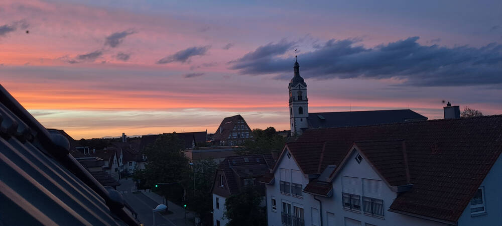 009_001 Das Bild zeigt eine Stadtansicht bei Sonnenuntergang mit einer Kirche und mehreren Gebäuden im Vordergrund, während der Himmel in bunten Pastelltönen gefärbt ist.