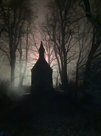 010_001 Eine kleine Kapelle steht im Vordergrund einer nebligen Nachtlandschaft, umgeben von kahlen Bäumen, während diffuses Licht den Hintergrund erhellt.