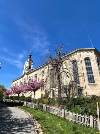 013_002 Eine große Kirche mit einem Kirchturm steht neben blühenden rosa Bäumen unter einem klaren blauen Himmel.