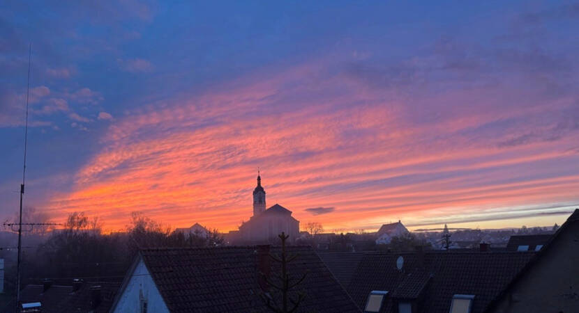 020_002 Ein farbenprächtiger Sonnenuntergang mit orange- und violett-gefärbtem Himmel erstreckt sich über den Dächern einer Stadt mit einer Kirche im Hintergrund.