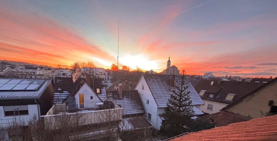 020_003 Ein farbenprächtiger Sonnenuntergang mit orange- und violett-gefärbtem Himmel erstreckt sich über den Dächern einer Stadt mit einer Kirche im Hintergrund.