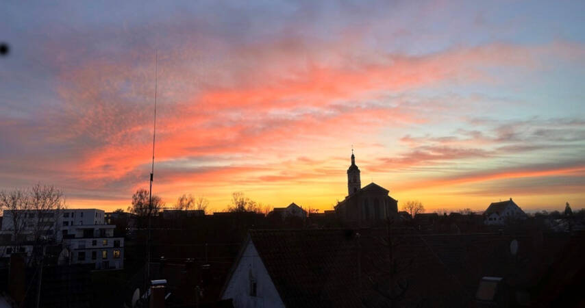 020_004 Ein farbenprächtiger Sonnenuntergang mit orange- und violett-gefärbtem Himmel erstreckt sich über den Dächern einer Stadt mit einer Kirche im Hintergrund.