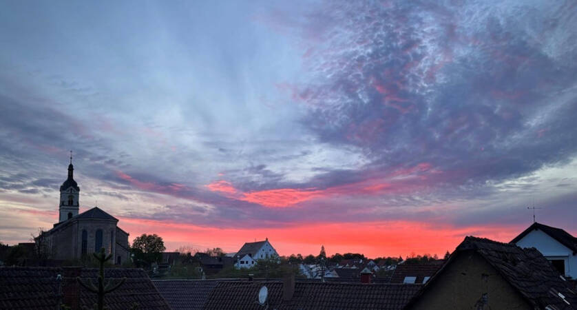 020_005 Ein farbenprächtiger Sonnenuntergang mit orange- und violett-gefärbtem Himmel erstreckt sich über den Dächern einer Stadt mit einer Kirche im Hintergrund.
