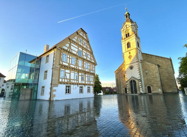 023_004 Ein historisches Fachwerkhaus steht neben einer großen Kirche auf einem mit Kopfsteinpflaster ausgelegten Platz unter klarem, blauem Himmel.
