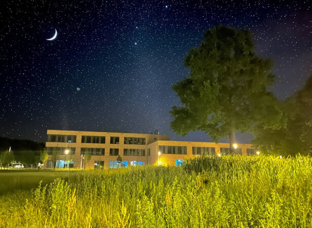 024_005 Ein modernes Gebäude steht bei Nacht unter einem klaren Sternenhimmel mit einer leuchtenden Mondsichel und einem großen Baum im Vordergrund.