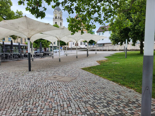 026_003 Einladende Atmosphäre auf einem beschirmten Platz mit Café, im Hintergrund stehen historische Gebäude und eine Kirche