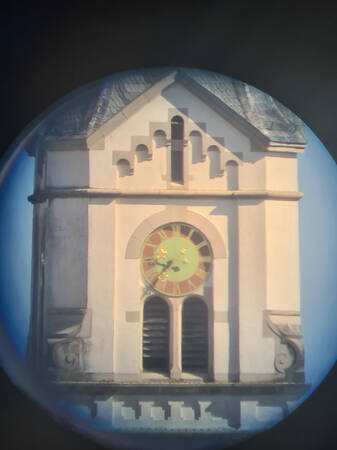 030_001 Das Bild zeigt ein Gebäude mit einem großen Zifferblatt und römischen Ziffern, das in einem steinernen, dreieckigen Giebel eingebettet ist.