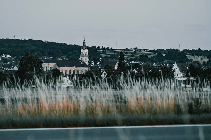 031_002 Im Vordergrund sind unscharfe Gräser und im Hintergrund eine Kirche und Häuserlandschaft vor einem bewaldeten Hügel zu sehen.