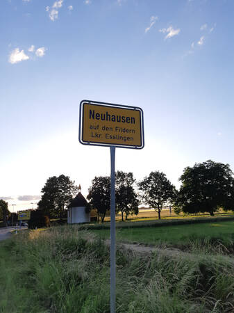 034_002 Ein gelbes Ortsschild mit der Aufschrift "Neuhausen auf den Fildern Lkr. Esslingen" steht am Straßenrand in einer ländlichen Umgebung mit Bäumen im Hintergrund.
