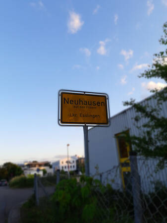 034_003 Ein gelbes Ortsschild mit der Aufschrift "Neuhausen auf den Fildern Lkr. Esslingen" steht am Straßenrand mit Bäumen und einem Gewerbebau im Hintergrund.