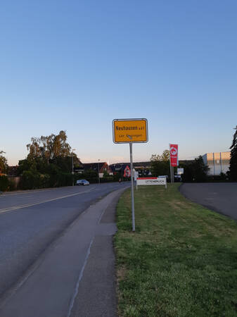 034_004 Ein gelbes Ortsschild mit der Aufschrift "Neuhausen auf den Fildern Lkr. Esslingen" steht am Straßenrand in einer ländlichen Umgebung.