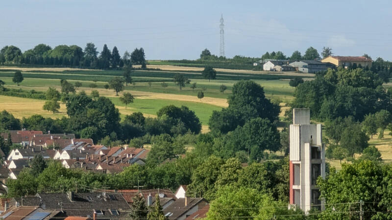 037_001 Das Bild zeigt eine ländliche Landschaft mit Feldern, Bäumen und Häusern im Vordergrund, während am Horizont Strommasten und Gebäude erkennbar sind.