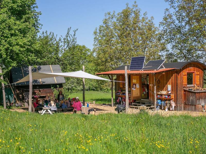 Eine Gruppe von Menschen sitzt unter einem Sonnensegel neben einer Holzhütte mit Solarpanelen, umgeben von Bäumen und einer grünen Wiese.