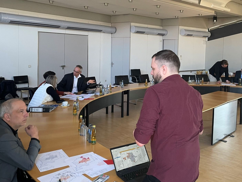 Ein Konferenzraum mit mehreren Personen, die an einem großen, ovalen Tisch arbeiten, mit Laptops, Karten und Getränkeflaschen.