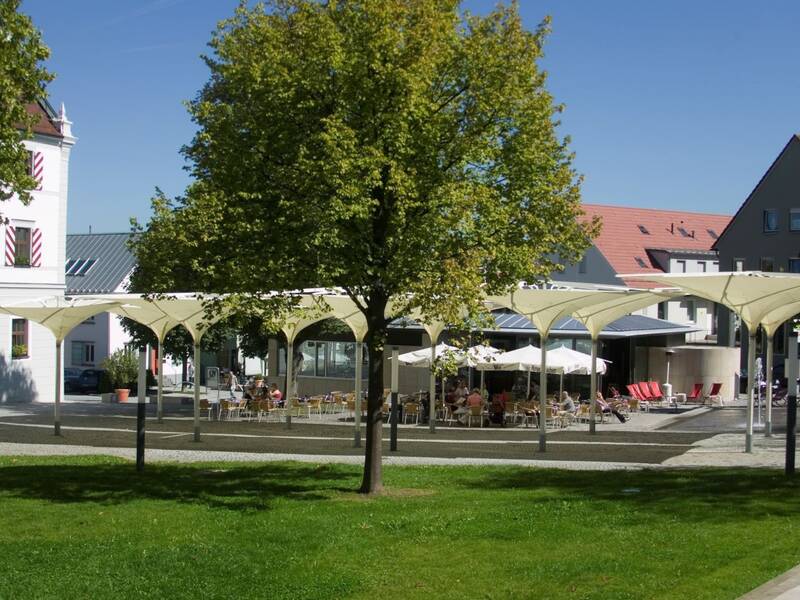Ein öffentlicher Platz mit modernen, weißen Sonnenschirmen, umgeben von Bäumen und Gebäuden mit rot-braunen Dächern.