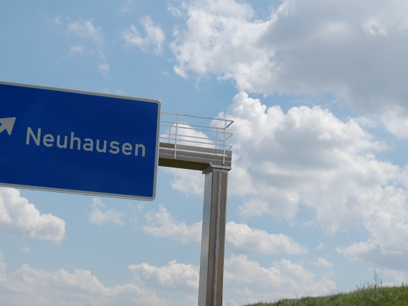 Ein blaues Straßenschild mit einem weißen Pfeil weist in Richtung "Neuhausen" vor einem himmelblauen Hintergrund mit weißen Wolken.