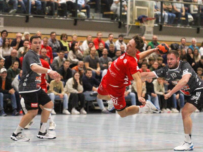 Ein Handballspieler in rotem Trikot springt dynamisch durch die Luft, während die gegnerischen Spieler in dunklen Trikots ihn verteidigen, und im Hintergrund füllen Zuschauer die Tribünen.