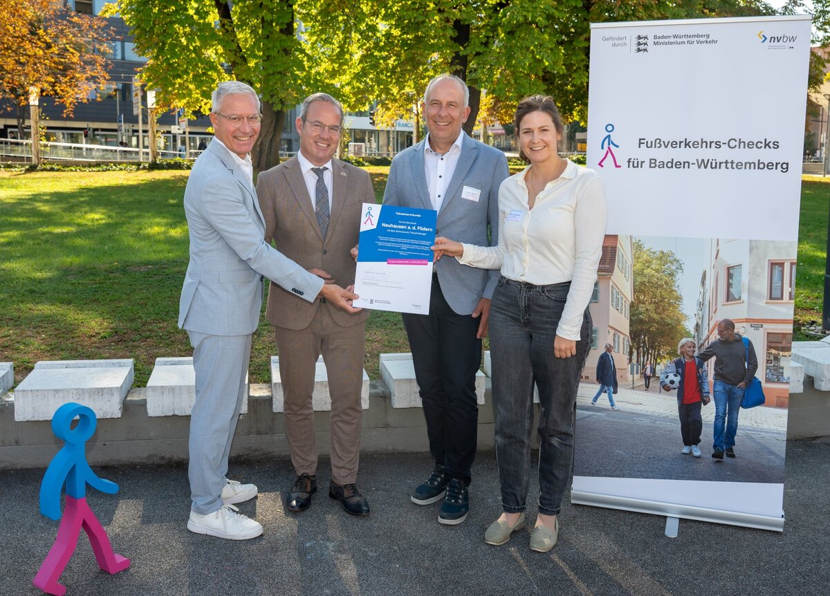 Vier Personen stehen lächelnd draußen und halten zusammen ein Zertifikat vor einem Banner mit der Aufschrift "Fußverkehrs-Checks für Baden-Württemberg".