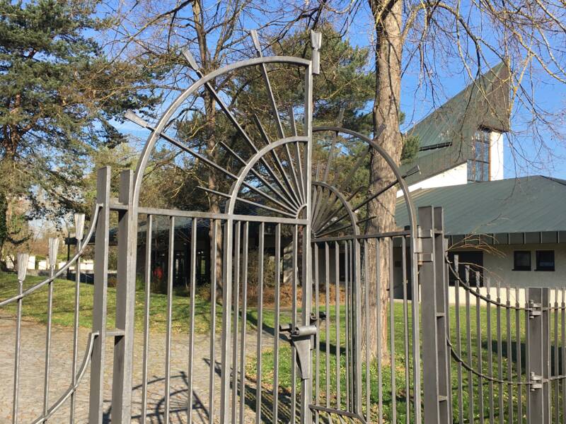 Ein schmiedeeisernes Tor mit geschwungenem Design steht offen auf einem Kopfsteinpflasterweg, umgeben von kahlen Bäumen, mit einem modernen Gebäude im Hintergrund bei sonnigem Wetter.