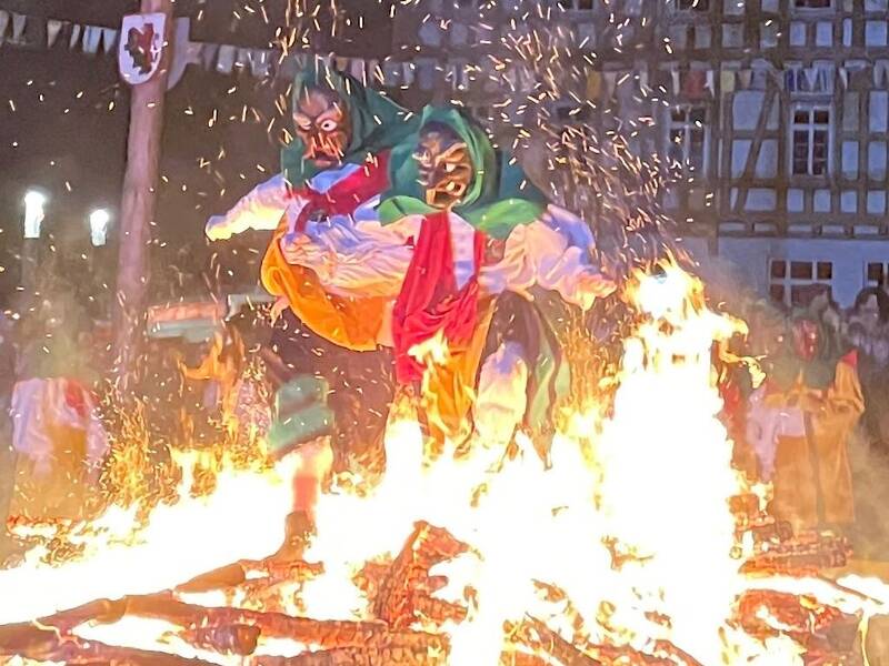 Zwei Personen in bunten, traditionellen Kostümen springen über ein großes Lagerfeuer bei einer nächtlichen Veranstaltung.